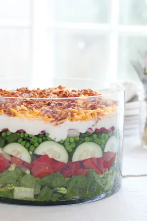 7-layer salad in a trifle dish with romaine, tomatoes, cucumbers, peas, radishes, sour cream dressing, cheddar, and bacon
