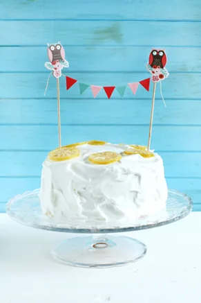 Paula Deen 7-minute frosting swirled on a white layer cake topped with candied lemon slices and an owl bunting on a glass cake stand