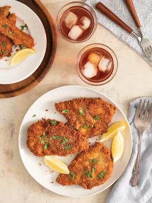 Paula Deen air fryer crispy fried chicken cutlets with lemon wedges and chopped parsley on a white plate beside two glasses of iced tea