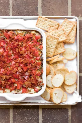 Paula Deen Backfield BLT dip in a white baking dish topped with chopped tomatoes and crumbled bacon, served with crackers and toasted bread rounds