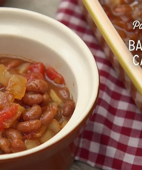 Paula Deen baked bean casserole with diced tomatoes and onion served in a small ceramic bowl beside a yellow baking dish
