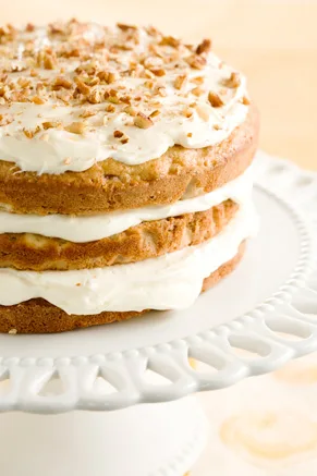 Paula Deen banana nut cake with cream cheese frosting, three layers topped with chopped pecans on a white cake stand