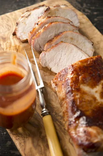 Sliced Paula Deen barbecue pork roast on a wooden cutting board with a carving fork and a jar of BBQ sauce
