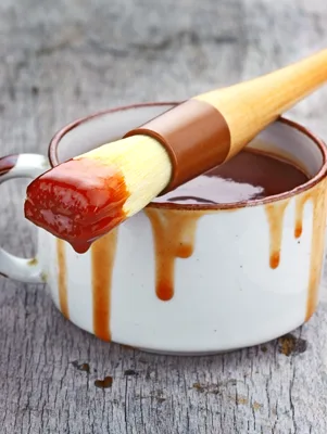 Paula Deen barbecue sauce in a white speckled mug with a basting brush coated in thick red-brown sauce resting on the rim