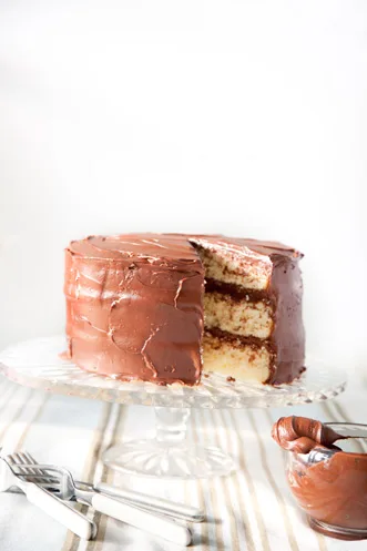 Paula Deen Basic 1-2-3-4 cake, a three-layer yellow cake with chocolate frosting and one slice removed on a glass cake stand