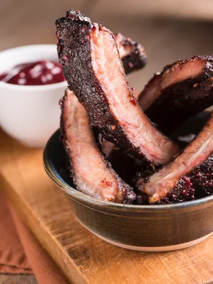Paula Deen BBQ baby back ribs with a dark glazed crust stacked in a bowl next to a small dish of BBQ sauce on a wooden board