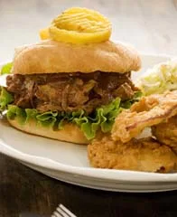 Paula Deen BBQ pork sandwich with shredded pork, lettuce and pickles on a seeded bun next to coleslaw and fried chicken on a white plate