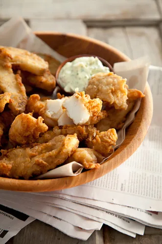 Paula Deen beer battered fish and chips piled in a wooden bowl on newspaper with a small dish of tartar sauce