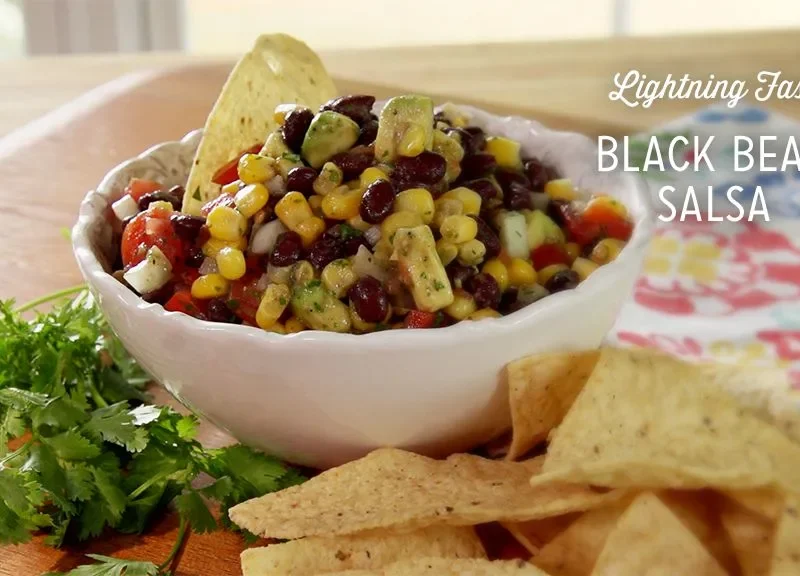 Paula Deen black bean salsa in a white scalloped bowl with a tortilla chip, surrounded by chips and fresh cilantro