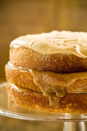 Paula Deen Bobby's Caramel Cake with two golden layers, caramel filling dripping from the middle and thick caramel frosting on top, on a glass cake stand