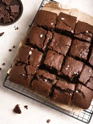 Paula Deen brownie cake cut into squares on a wire rack, topped with flaky sea salt, with chocolate pieces in a bowl nearby