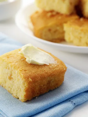 Paula Deen buttermilk cornbread square topped with a pat of butter on a blue napkin, with more squares on a plate behind