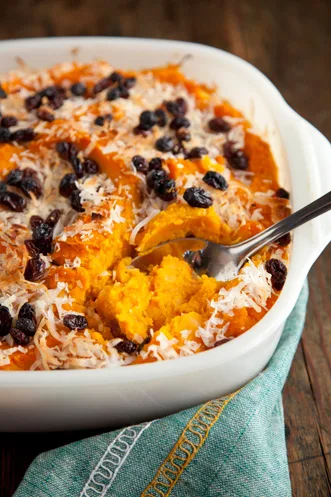Paula Deen butternut squash casserole in a white baking dish topped with raisins and flaked coconut, with a spoon scooping a serving