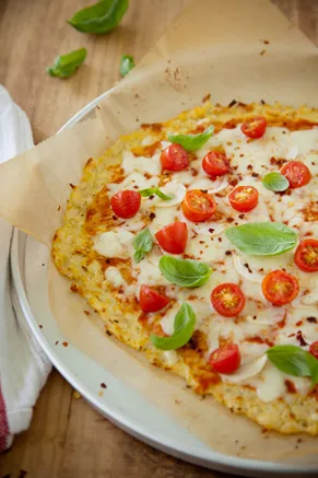 Paula Deen cauliflower crust pizza topped with melted mozzarella, halved grape tomatoes, fresh basil and red pepper flakes on parchment paper