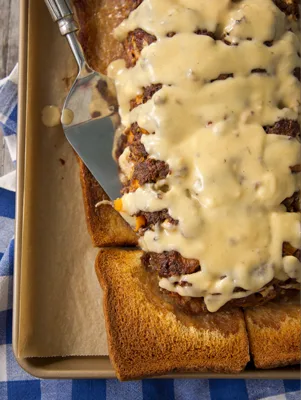 Paula Deen cheeseburger meatloaf and sauce sliced on a baking sheet over toasted bread, covered in creamy cheddar sauce