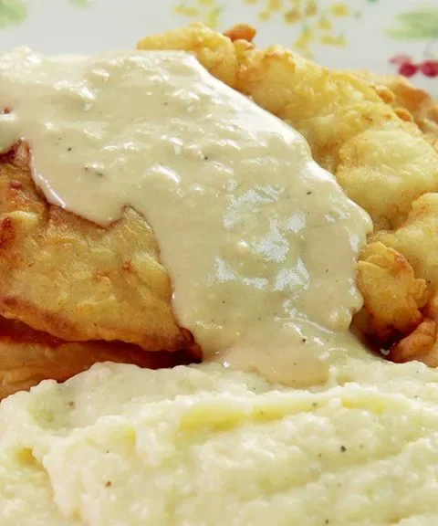 Paula Deen cheesy grits served in a creamy mound on a floral plate beside golden fried chicken topped with gravy