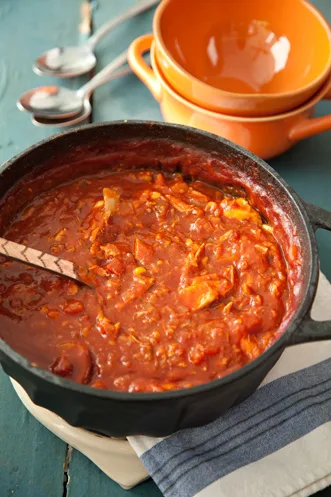 Paula Deen chicken Brunswick stew simmering in a black cast iron pot with a wooden spoon, orange bowls stacked behind it