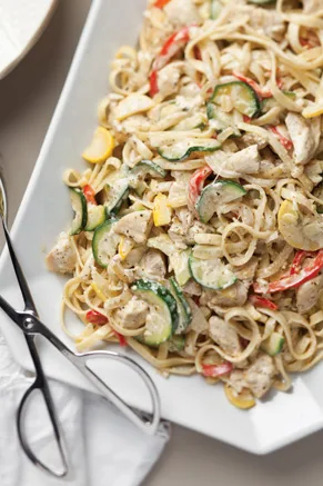 Paula Deen chicken pesto pasta with fettuccine, chicken pieces, zucchini slices and red bell pepper on a white platter with tongs
