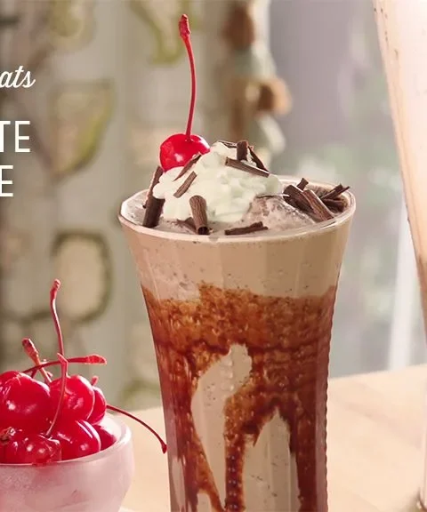 Paula Deen chocolate brownie shake in a tall glass with chocolate syrup streaks, whipped cream, shaved chocolate and a maraschino cherry