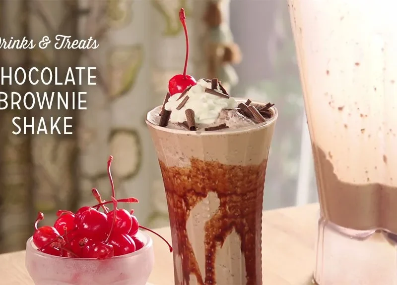 Paula Deen chocolate brownie shake in a tall glass with chocolate syrup streaks, whipped cream, shaved chocolate and a maraschino cherry