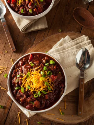 Paula Deen chocolate chili in a white bowl topped with shredded cheese and sliced green onions on a wooden table