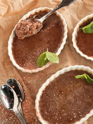 Paula Deen chocolate creme brûlée in white fluted ramekins with a sugar crust, fresh mint leaves and a spoon lifting out a scoop of custard