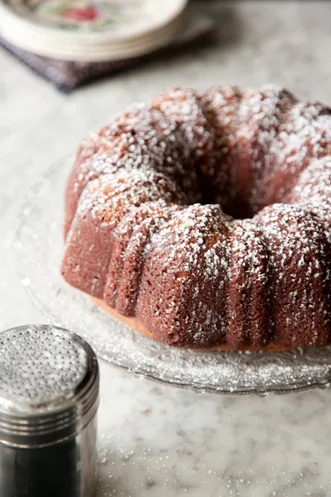 Paula Deen chocolate sour cream pound cake dusted with powdered sugar on a glass cake stand with a sugar shaker nearby