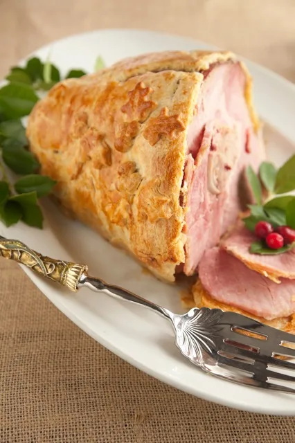 Paula Deen Christmas ham wrapped in golden crescent dough, sliced on a white platter with holly leaves and red berries