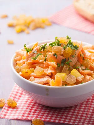 Paula Deen corn and carrot salad with golden raisins and fresh parsley in a white bowl on a red checkered cloth