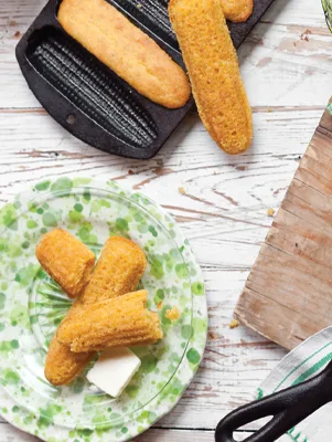 Paula Deen corn sticks golden brown on a green patterned plate with butter next to a cast-iron corn stick pan