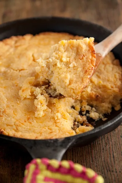 Paula Deen corny cornbread in a cast iron skillet with a wooden spoon lifting out a golden, moist slice