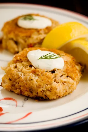 Paula Deen crab cakes with lemon-dill sauce, golden brown patties topped with a dollop of dill sauce and lemon wedges on a white plate