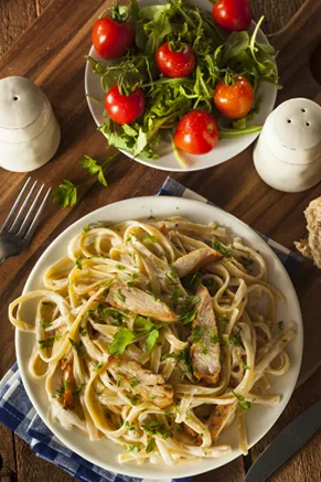 Paula Deen fettuccine chicken salad with baked chicken strips and fresh herbs on a white plate beside a bowl of arugula and cherry tomatoes