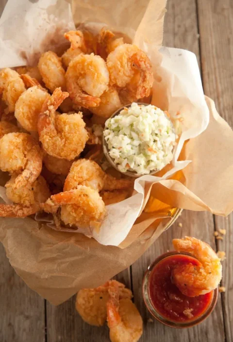 Paula Deen fried shrimp piled in parchment paper with a cup of coleslaw and cocktail sauce on a wooden table