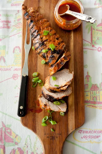 Paula Deen grilled pork tenderloin sliced on a wooden board with sliced scallions and a jar of glaze