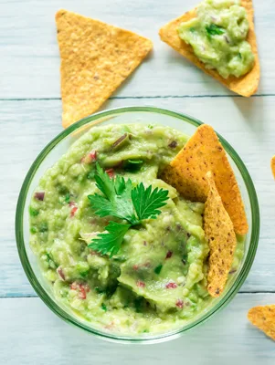 Paula Deen guacamole in a glass bowl with a cilantro garnish and tortilla chips on a light blue wooden table
