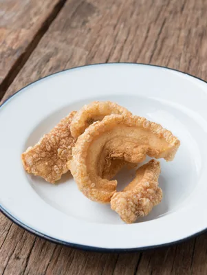 Paula Deen home made pork rinds, golden crispy puffed pieces piled on a white enamel plate with a dark rim