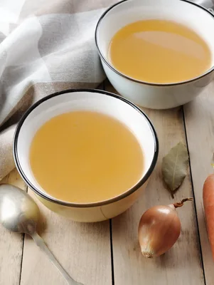 Two bowls of Paula Deen homemade chicken stock, golden and clear, beside an onion, bay leaf and carrot on a wooden table