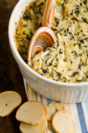 Paula Deen hot spinach-artichoke dip baked in a white dish with a colorful wooden spoon and sliced bread beside it