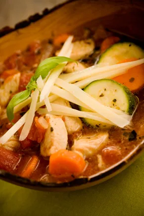 Paula Deen Italian chicken and vegetable soup with chicken pieces, zucchini, carrots and tomatoes topped with shaved Parmesan and a basil leaf in a wooden bowl