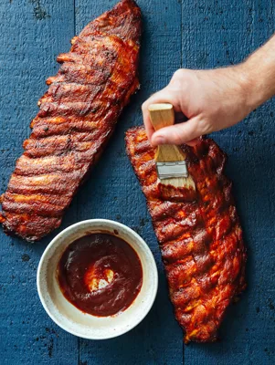 Paula Deen Jamie's BBQ Sauce in a white bowl beside two glazed racks of ribs on a blue wooden table with a basting brush