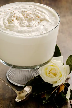 Paula Deen Mama's eggnog in a glass pedestal bowl topped with nutmeg beside a white rose and a spoon