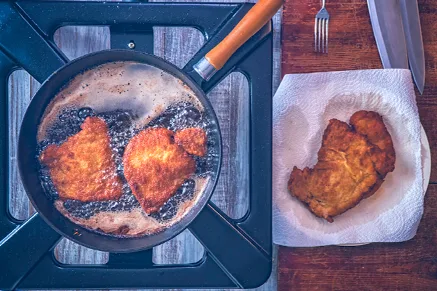 Paula Deen Mama's fried pork chops frying golden brown in a black skillet next to a paper towel-lined plate with a finished chop
