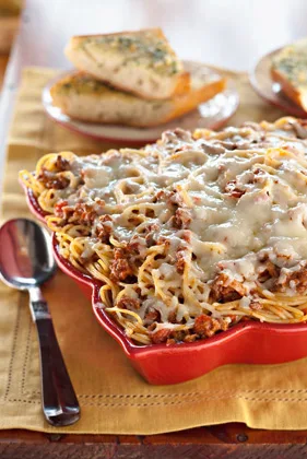Paula Deen Mama's spaghetti casserole with melted mozzarella in a red baking dish with garlic herb bread on a plate behind