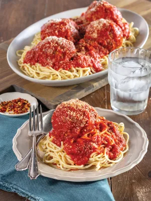 Paula Deen meatballs topped with red sauce and grated Parmesan over spaghetti on a plate with a serving platter behind