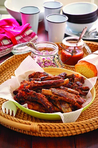 Paula Deen Memphis dry rub ribs piled in a green bowl lined with parchment on a woven tray with cornbread and coleslaw