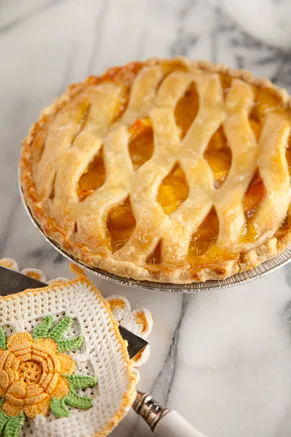 Paula Deen Nita's Secret Peach Pie with a golden lattice crust on a marble counter beside a crocheted pot holder