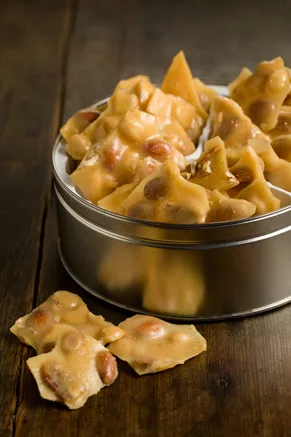 Paula Deen nutty brittle pieces with almonds and pecans piled in a round metal tin on a dark wooden table