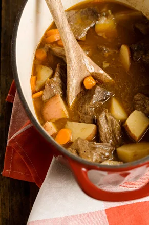 Paula Deen old-time Southern beef stew with chunks of beef, red potatoes and carrots in a red Dutch oven with a wooden spoon