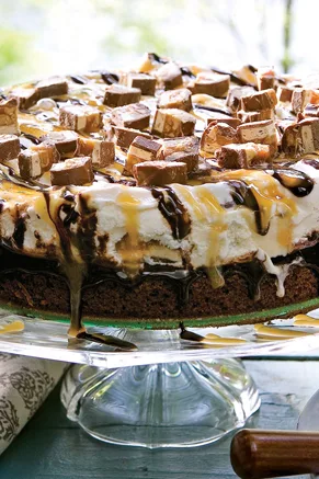 Paula Deen ooey gooey chocolate ice cream cake with chopped Snickers pieces and chocolate and caramel drizzle on a glass cake stand